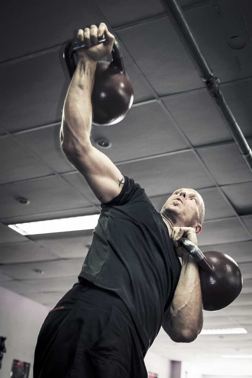 En person i svart t-shirt utför en pressövning med kettlebell över huvudet i ett gym.