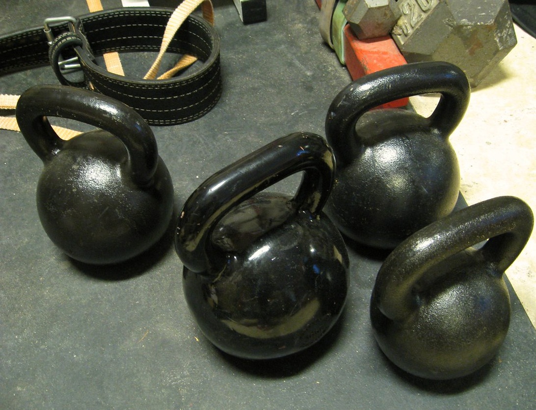 En samling svarta kettlebells i olika storlekar på ett gymgolv, med ett lyftarbälte i bakgrunden.