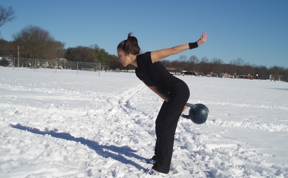 En person i helsvarta träningskläder utför en kettlebellövning i ett snötäckt fält, vilket illustrerar sportanpassad träning för skidåkning.